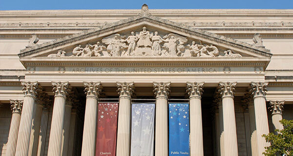 View of the top of National Archives building in Washington DC made up of greek style columns and a pediment with greek figures and the words 'ARCHIVES OF THE UNITED STATES OF AMERICA'