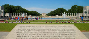 View of Washington DC World War II Memorial featuring a large stone inscription in foreground, a large fountain in the middle, and large columns surrounding fountain with flower bouquets surrounding monument and the Lincoln Memorial in background