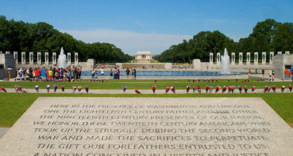 View of Washington DC World War II Memorial featuring a large stone inscription in foreground, a large fountain in the middle, and large columns surrounding fountain with flower bouquets surrounding monument and the Lincoln Memorial in background