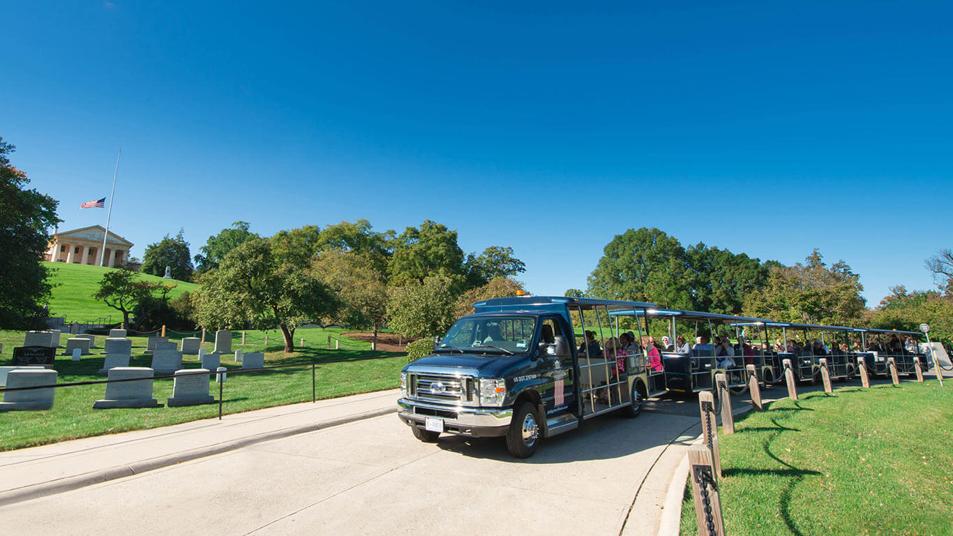 Arlington Cemetery Tour vehicle filled with guests driving past head stones and Arlington House and US flag sitting on a hill in the background