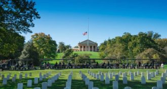 Arlington National Cemetery Tours vehicle driving past rows of gravestones in the foreground and Arlington House and US flag on pole in the background