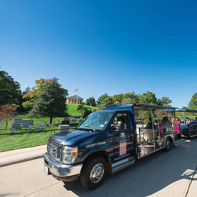 Arlington Cemetery Tour vehicle filled with guests driving past head stones and Arlington House and US flag sitting on a hill in the background